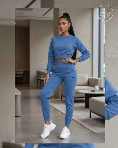 A woman modeling a matching blue two-piece activewear set consisting of a long-sleeve crop top and jogger pants, styled with white sneakers in a modern indoor setting. A brand logo for 'Divine Inspirings' is visible in the top right corner.