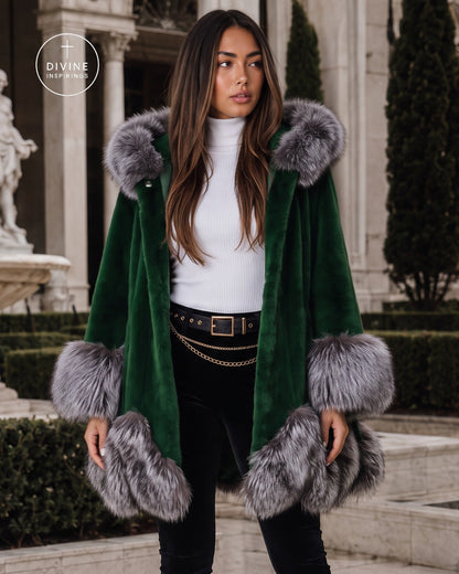 A woman wearing a dark green velvet coat with gray fur trim, styled with a white turtleneck, black pants, and a gold chain belt, standing in front of a grand classical building.