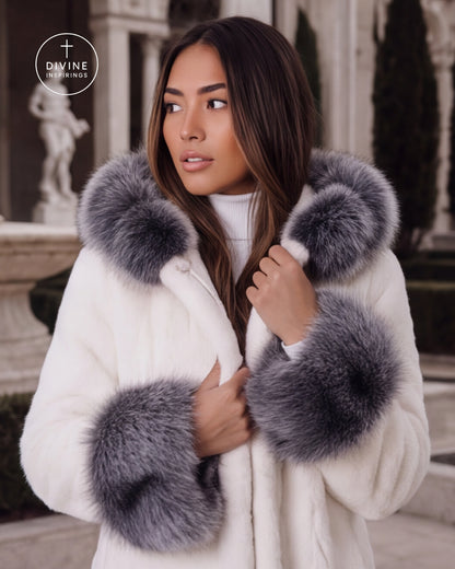 A woman wearing a white faux fur coat with large gray fur trim on the collar and cuffs, standing outdoors in front of a classical building. A circular 'DIVINE INSPIRINGS' logo is visible in the top left corner.