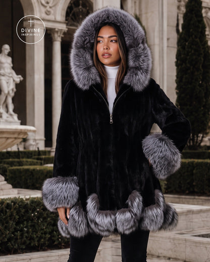 A woman wearing a black faux fur coat with a large gray fur-trimmed hood, cuffs, and hem, standing outdoors in front of a classical building with columns and hedges. A circular 'DIVINE INSPIRINGS' logo is visible in the top left corner.