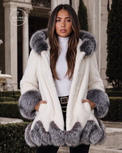 A woman wearing a cream faux fur coat with gray fur trim on the collar, cuffs, and hem, standing outdoors in front of a classical building with columns.