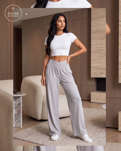 A woman modeling a white short-sleeve crop top and light gray wide-leg pants in a modern indoor setting with wood paneling, a beige sofa, and a rug. The Divine Inspirings logo is visible in the top left corner.