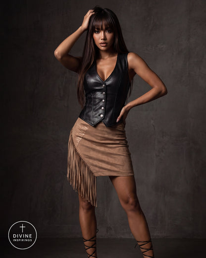 A woman modeling a black faux-leather vest corset and a brown fringe mini skirt against a dark textured studio backdrop. A Divine Inspirings logo is visible in the bottom-left corner.