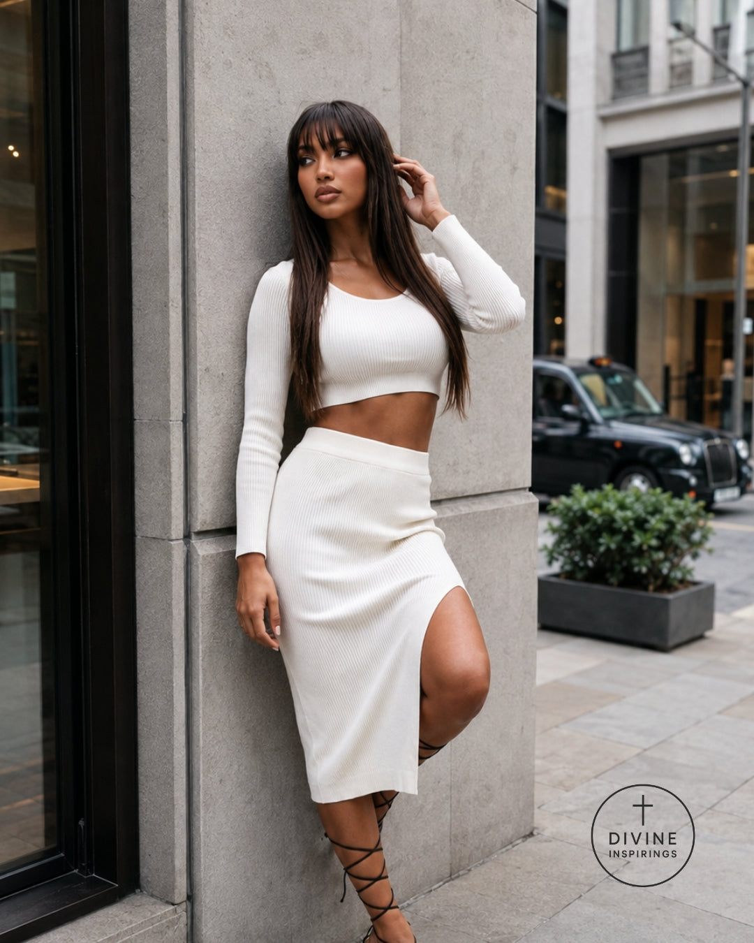 A woman posing in a white long-sleeve crop top and matching high-waisted midi skirt with a thigh-high slit, set against a modern concrete building exterior.