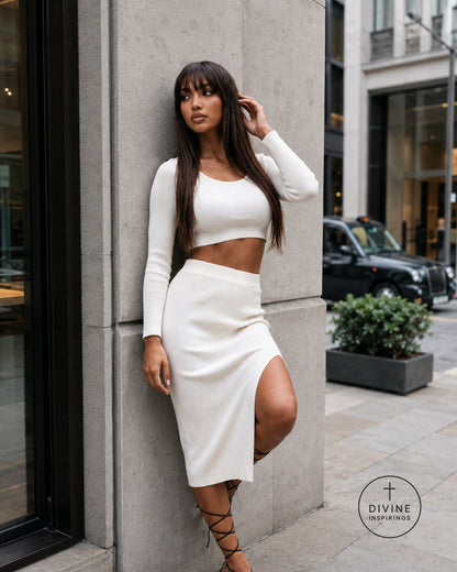 A woman posing in a white long-sleeve crop top and matching high-waisted midi skirt with a thigh-high slit, set against a modern concrete building exterior.