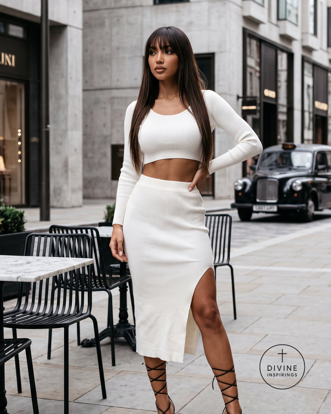 A woman stands on a city street wearing a matching ribbed knit two-piece outfit consisting of a white long-sleeve crop top and a high-slit midi skirt. The background features urban architecture, outdoor cafe tables, and a classic black cab.
