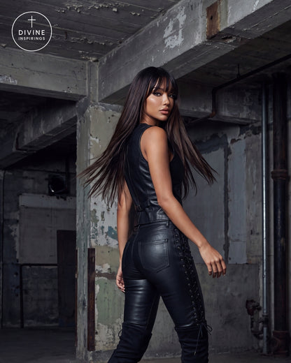 A woman with long dark hair models a black faux-leather sleeveless top and matching high-waisted lace-up pants in a gritty, industrial loft setting. The Divine Insprings logo is visible in the top-left corner.