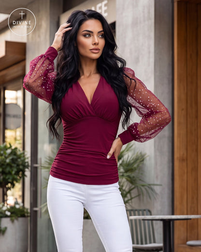 A woman modeling a deep burgundy long-sleeve top with sheer polka-dot puff sleeves, paired with white pants, standing in front of a modern building entrance.