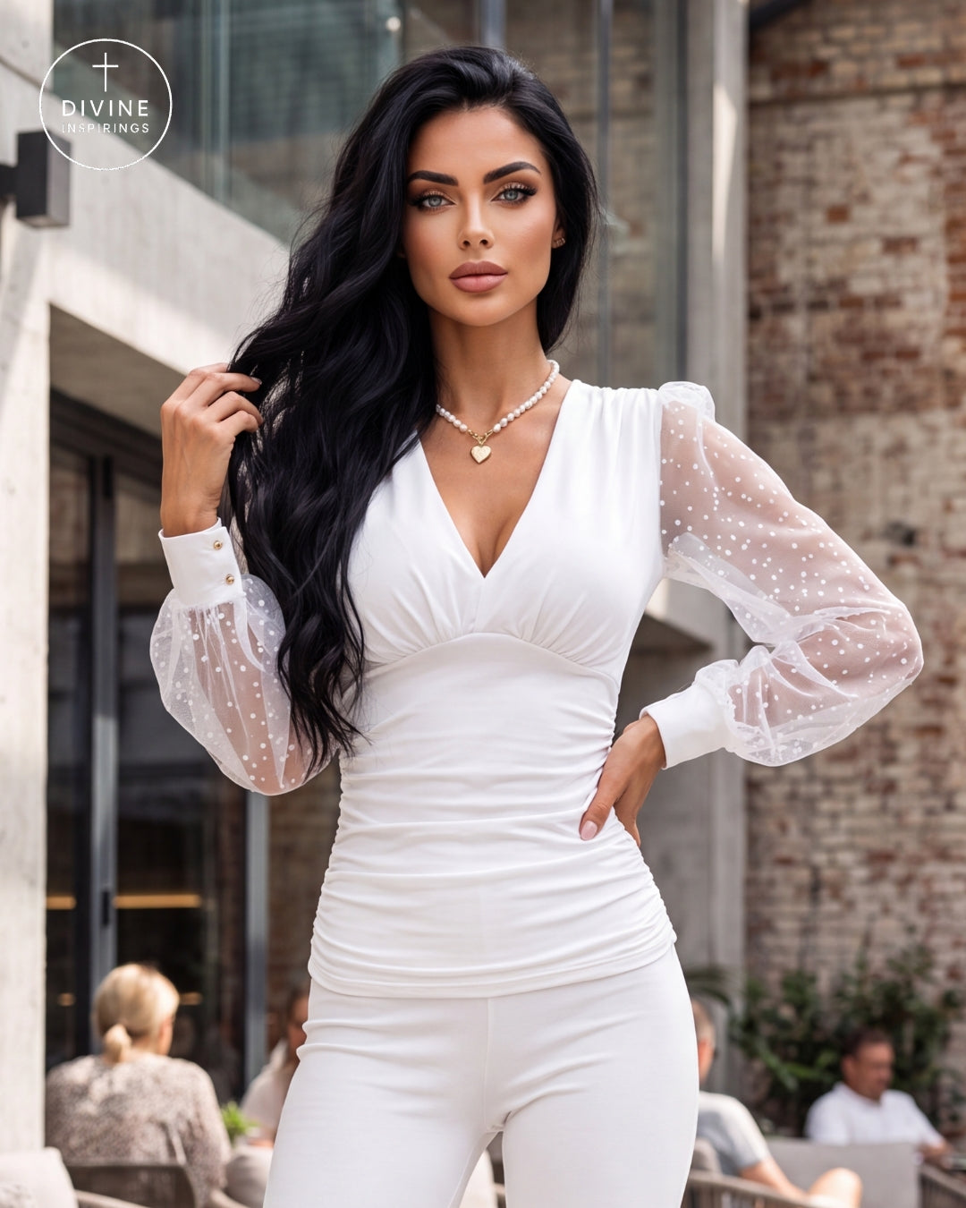 A woman modeling a white ruched bodysuit with sheer long sleeves featuring white polka dots, posed in front of a modern brick building.