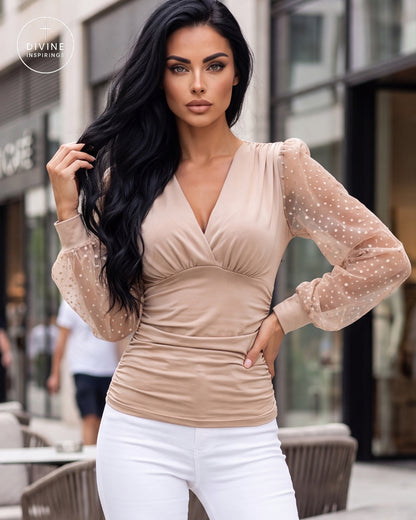 A woman modeling a beige long-sleeve top with sheer polka-dot sleeves, paired with white pants, in an outdoor urban setting. A circular 'DIVINE INSPIRINGS' watermark is visible in the top-left corner.