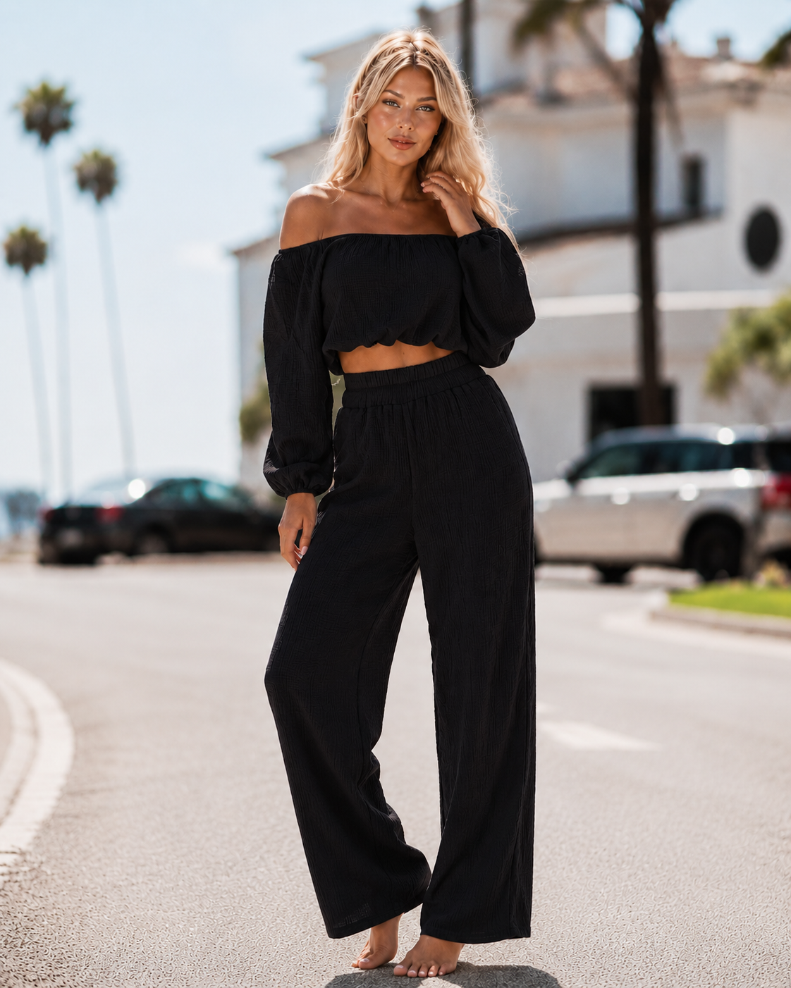 Woman in black off-shoulder top and wide-leg pants standing on a street with palm trees in the background.