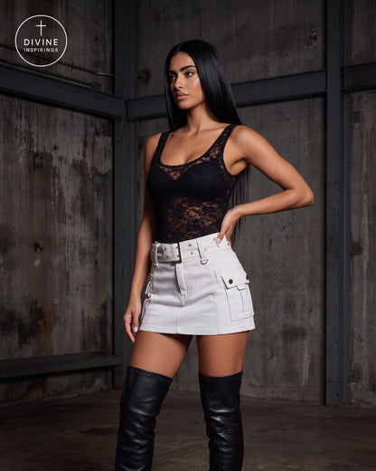 A woman modeling a black lace camisole top paired with a white cargo mini skirt and black over-the-knee boots, standing against a dark industrial metal wall. A Divine Inspirings logo is visible in the top-left corner.