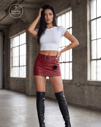 A woman stands in a concrete industrial loft wearing a white cropped short-sleeve top, a red faux-leather mini skirt with a black belt and pom-pom, and black over-the-knee boots. A circular 'DIVINE INSPIRINGS' logo is in the top-left corner.