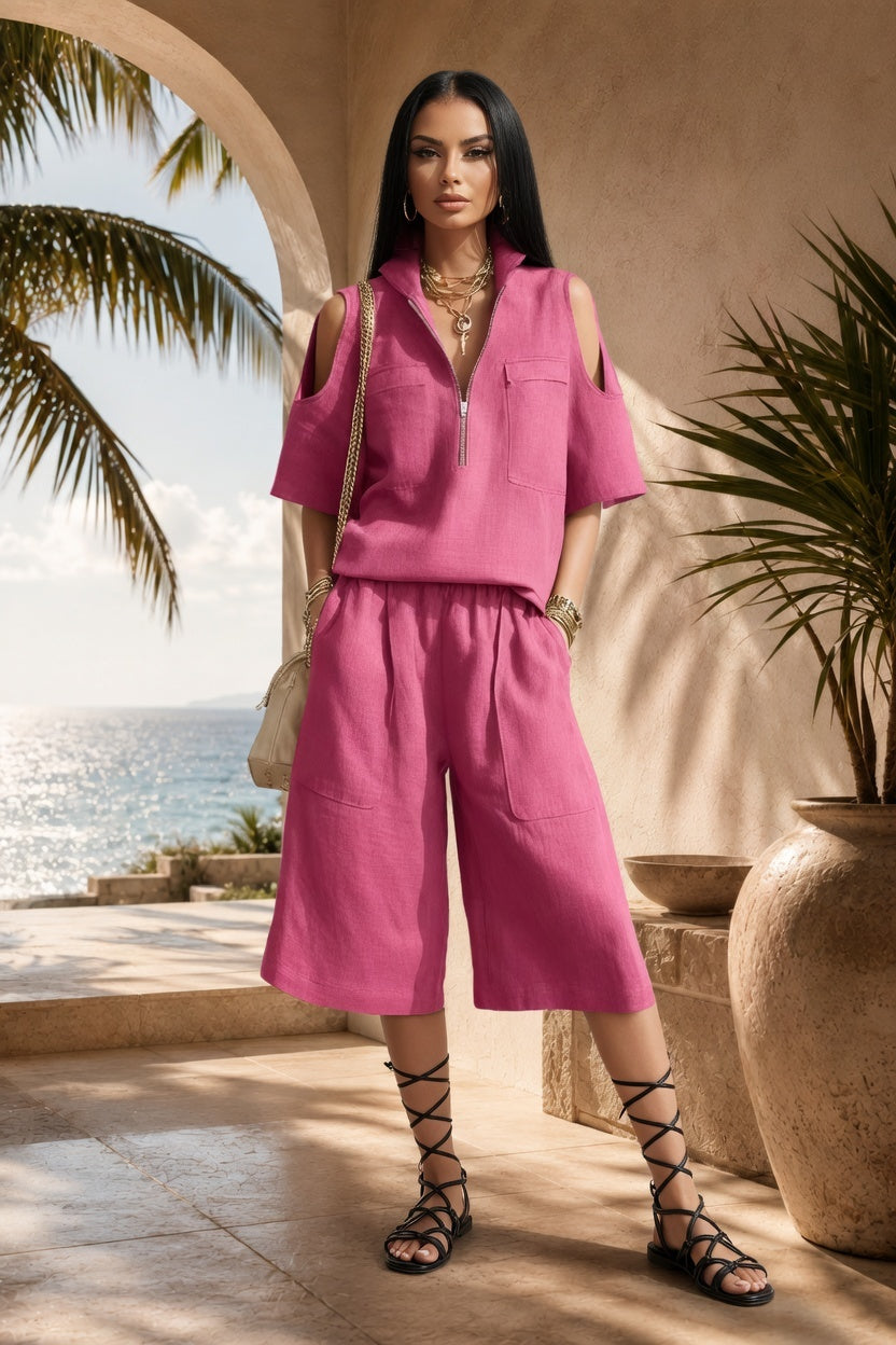 A woman stands outdoors in a sunlit architectural setting with a sea view behind her, wearing a bright pink two-piece outfit with a short-sleeve collared top and matching wide-leg cropped pants. She is styled with layered gold necklaces, a shoulder bag, and black lace-up sandals.