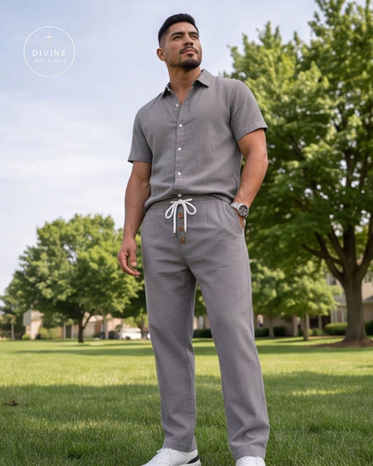 A man wearing a light gray short-sleeve button-up shirt and matching drawstring pants stands on a grassy park lawn with trees in the background. A circular 'DIVINE' logo watermark is visible in the top-left corner.