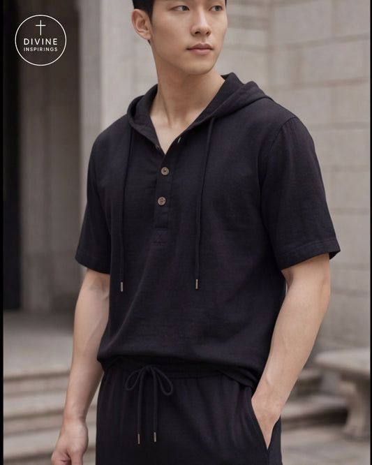 A man wearing a black short-sleeve hooded pullover with drawstrings and a button placket, paired with matching black drawstring pants, photographed outdoors against a blurred stone wall. A circular 'DIVINE INSPIRATION' logo appears in the top-left corner.