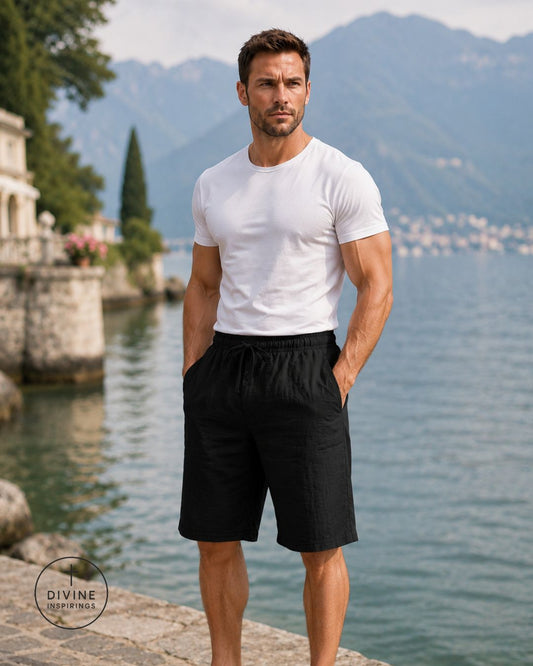 A man standing outdoors by a lake with mountains in the background, wearing a fitted white short-sleeve crew-neck t-shirt and black drawstring shorts. A circular 'DIVINE INSPIRINGS' watermark is visible in the lower-left corner.