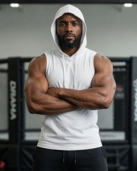 A muscular man wearing a white sleeveless hoodie and black sweatpants stands with arms crossed in a gym setting.