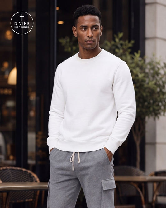 A man wearing a white long-sleeve crewneck sweatshirt and gray cargo jogger pants, standing in front of a modern building with glass windows and outdoor seating. A circular 'DIVINE INSPIRINGS' logo is visible in the top left corner.