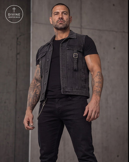 A muscular man with tattoos models a black denim vest over a black t-shirt and black jeans, standing against a gray concrete wall. The DIVINE INSPIRINGS logo is visible in the top left corner.