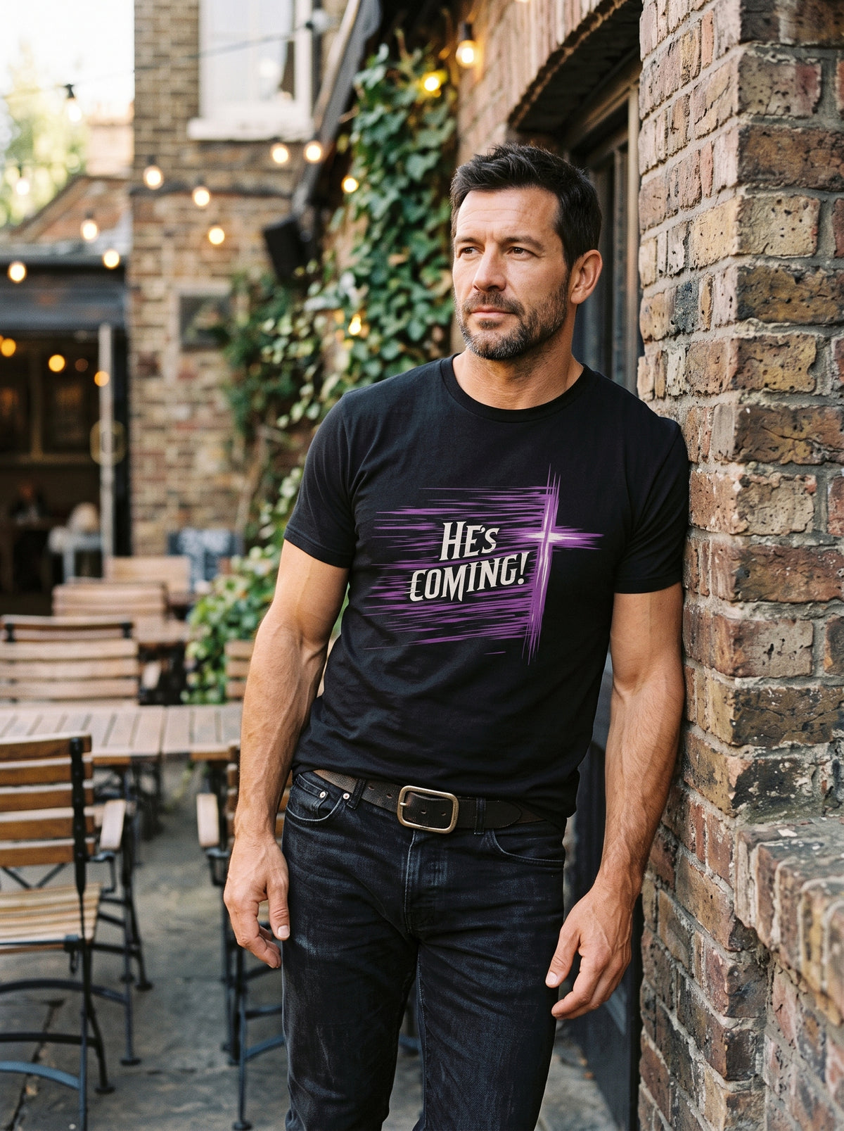 A man wearing a black graphic t-shirt with a purple and white 'HES COMING!' design, standing outdoors against a brick wall.