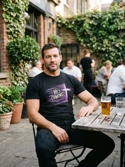 A man sitting at an outdoor patio table wearing a black graphic t-shirt with the text 'HES COMING!' in white and purple. The background shows a brick pub interior with green ivy, other patrons, and a glass of beer on the table.