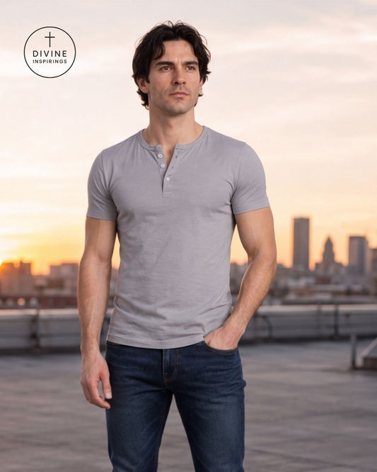 A man wearing a light gray short-sleeve henley shirt and dark blue jeans, standing on a rooftop at sunset with a blurred city skyline in the background. A circular Divine Inspirations logo is visible in the top-left corner.