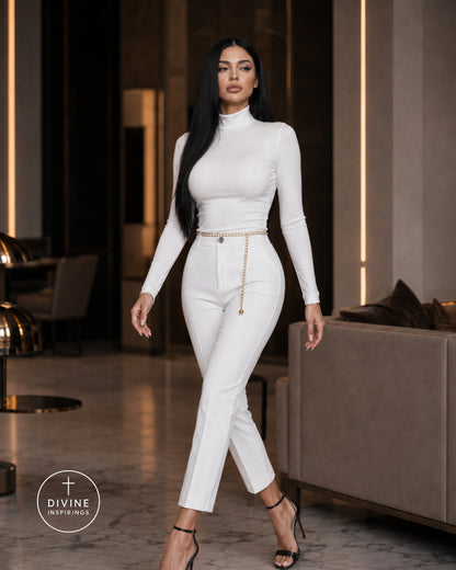 A woman modeling a white long-sleeve mock-neck top and matching white cropped trousers with a thin gold chain belt, standing in a modern indoor setting with a sofa and warm vertical light strips in the background.