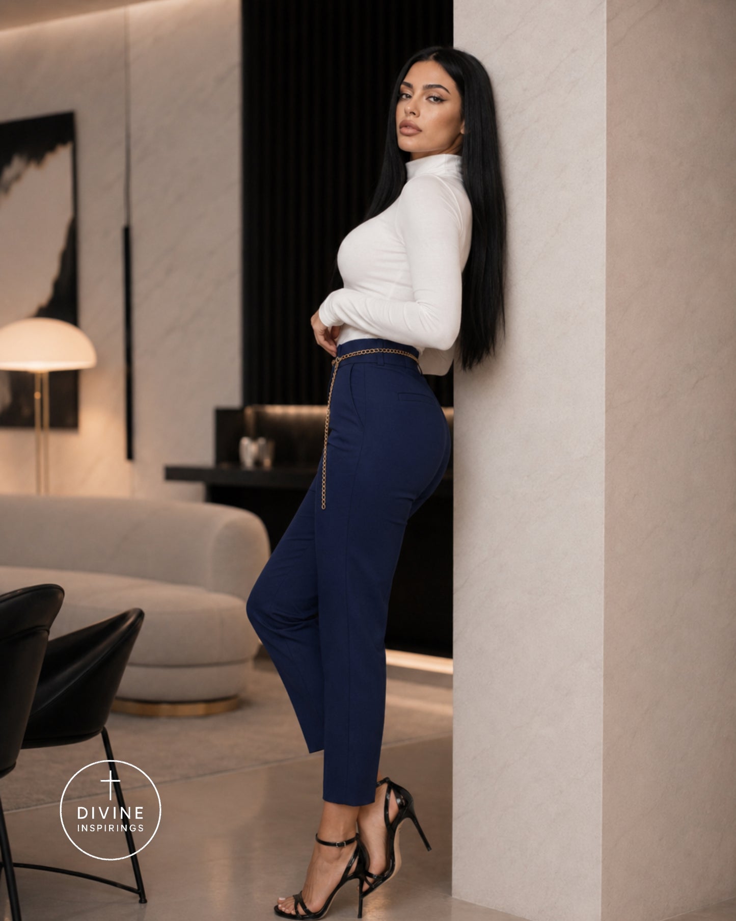 A woman posing in a modern interior, wearing a white mock-neck long-sleeve top and navy blue high-waisted cropped pants with a gold zipper detail at the waist, paired with black strappy heels. A 'DIVINE INSPIRINGS' watermark is visible in the bottom left corner.
