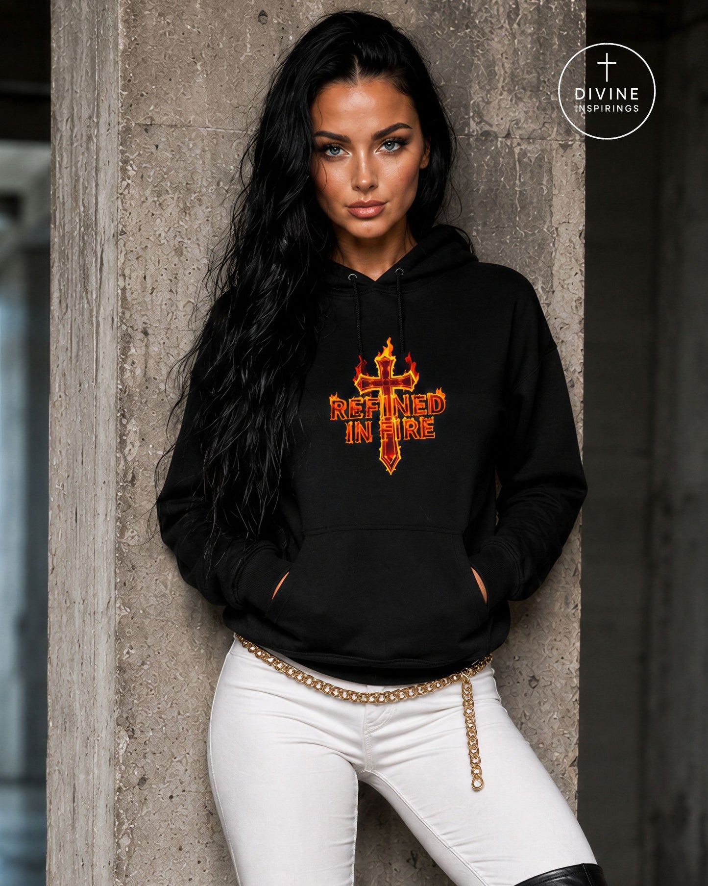 A woman wearing a black pullover hoodie with a red and orange graphic that reads 'REFINED IN FIRE' under a flaming cross, paired with white pants and a gold chain belt, leaning against a concrete pillar.