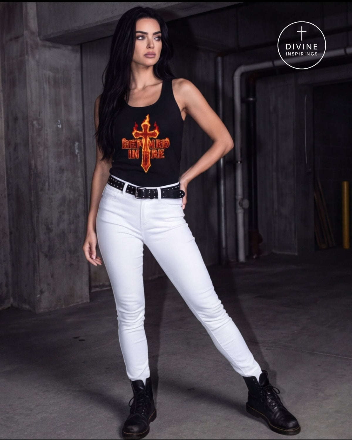 A woman wearing a black tank top with a large orange and yellow flame-cross graphic that says "Refined in Fire" on the chest, paired with white fitted pants and a gold chain belt, standing against a dark concrete wall. A 'DIVINE INSPIRINGS' logo is visible in the top right corner.