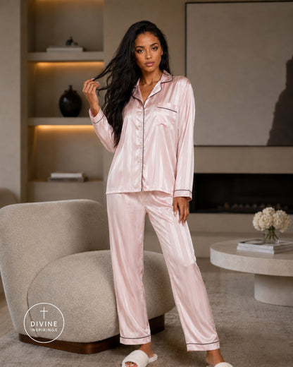 A woman modeling a light pink satin pajama set with matching pants in a modern living room setting. The pajama set features a long-sleeve button-up top and matching pants with subtle piping details.