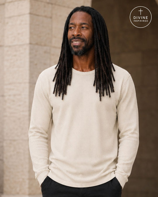 A man with long black dreadlocks wearing a cream-colored waffle-knit long-sleeve shirt and black pants, standing outdoors against a neutral stone wall. A circular 'Divine Inspirings' logo is visible in the top right corner.