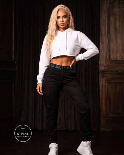 A woman modeling a white cropped long-sleeve hoodie and black cargo pants with a black studded belt, standing in front of a dark wood-paneled background. A circular 'DIVINE INSPIRINGS' logo is visible in the bottom-left corner.