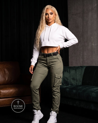 A woman posing in a white cropped hoodie and olive green cargo pants with white sneakers, set against a dark studio background with a brown leather and green velvet sofa.