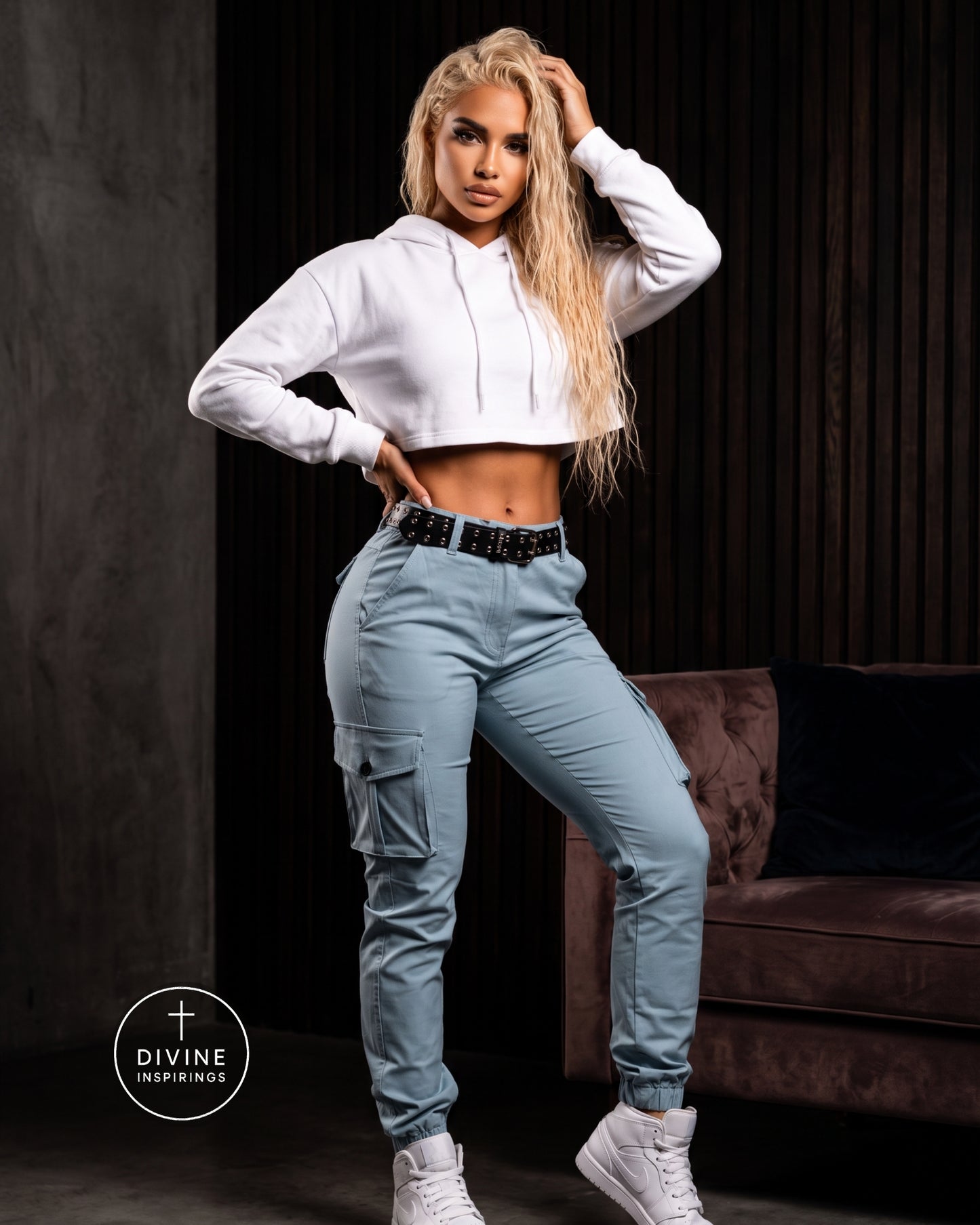 A woman posing in a white cropped hoodie and light blue cargo pants with a black studded belt, standing in a dark room with a brown velvet sofa in the background. A 'DIVINE INSPIRINGS' logo is visible in the bottom-left corner.