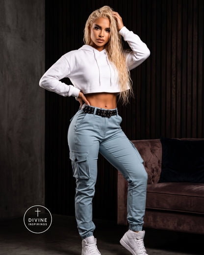 A woman posing in a white cropped hoodie and light blue cargo pants with a black studded belt, standing in a dark room with a brown velvet sofa in the background. A 'DIVINE INSPIRINGS' logo is visible in the bottom-left corner.