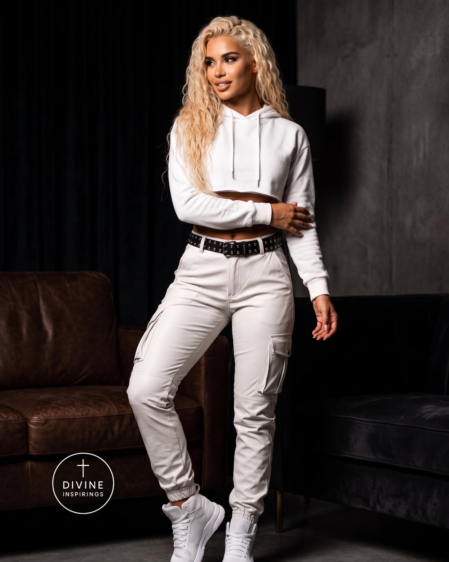 A woman modeling a white cropped hoodie and matching white cargo pants with a black studded belt, standing in a dark room with brown and black sofas. A circular 'DIVINE INSPIRINGS' logo is visible in the lower left corner.