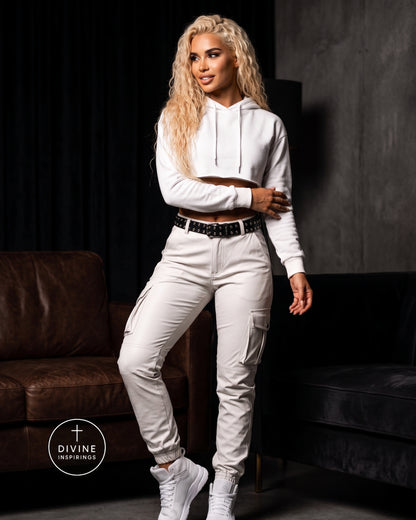 A woman modeling a white cropped hoodie and matching white cargo pants with a black studded belt, standing in a dark room with brown and black sofas. A circular 'DIVINE INSPIRINGS' logo is visible in the lower left corner.