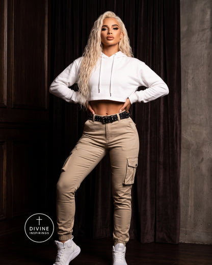 A blonde model poses in a white cropped hoodie and beige cargo jogger pants with white sneakers, standing against a dark curtain background. A circular 'DIVINE INSPIRINGS' logo is visible in the bottom-left corner.