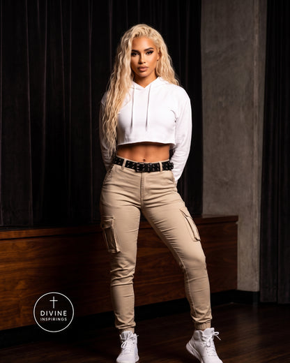 A woman modeling beige cargo pants with a black belt, styled with a white cropped hoodie and white sneakers, standing in front of a dark wood-paneled wall.