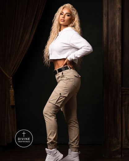 A blonde model poses in a white cropped hoodie and beige cargo pants with white sneakers, set against a dark background with curtains.