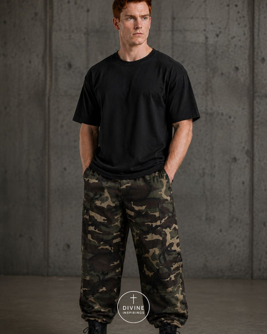 A man wearing a black crewneck t-shirt and camouflage cargo pants stands against a textured concrete wall. A circular logo for 'DIVINE INSPIRED' is visible at the bottom center.