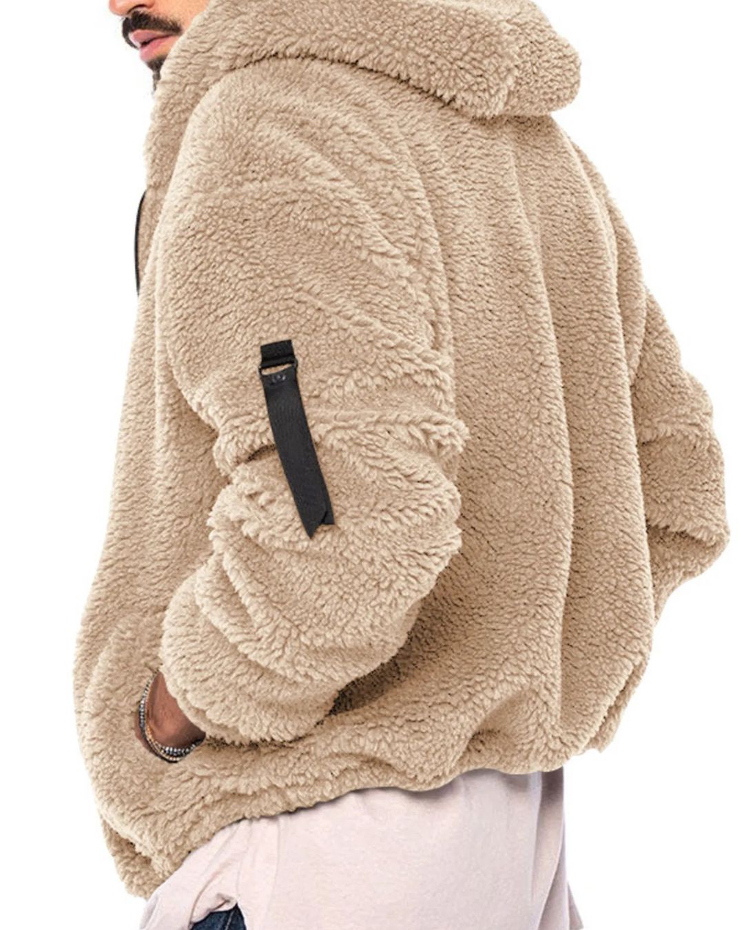 A close-up side view of a person wearing a beige sherpa fleece hooded jacket with a black sleeve strap detail, paired with light pink pants against a white background.