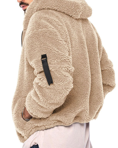 A close-up side view of a person wearing a beige sherpa fleece hooded jacket with a black sleeve strap detail, paired with light pink pants against a white background.