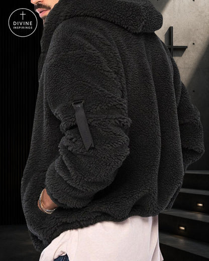 A close-up of a person wearing a dark charcoal gray sherpa fleece hooded jacket with a black strap detail on the sleeve, paired with white pants. The DIVINE INSPIRINGS logo is visible in the top-left corner.