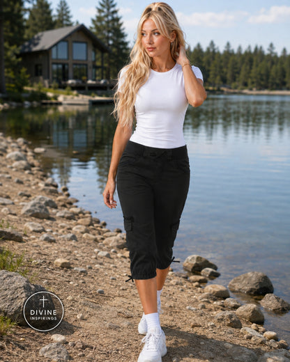A woman walking along a rocky lakeshore wearing a fitted white short-sleeve top and black cropped cargo pants. A house and pine trees are visible in the background, and a circular 'DIVINE DESIGNERS' logo appears in the lower-left corner.