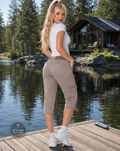 A woman stands on a wooden dock by a lake wearing loose-fit cropped cargo pants in a light taupe/khaki color, paired with a white t-shirt and white sneakers. A cabin and pine trees are visible in the background.
