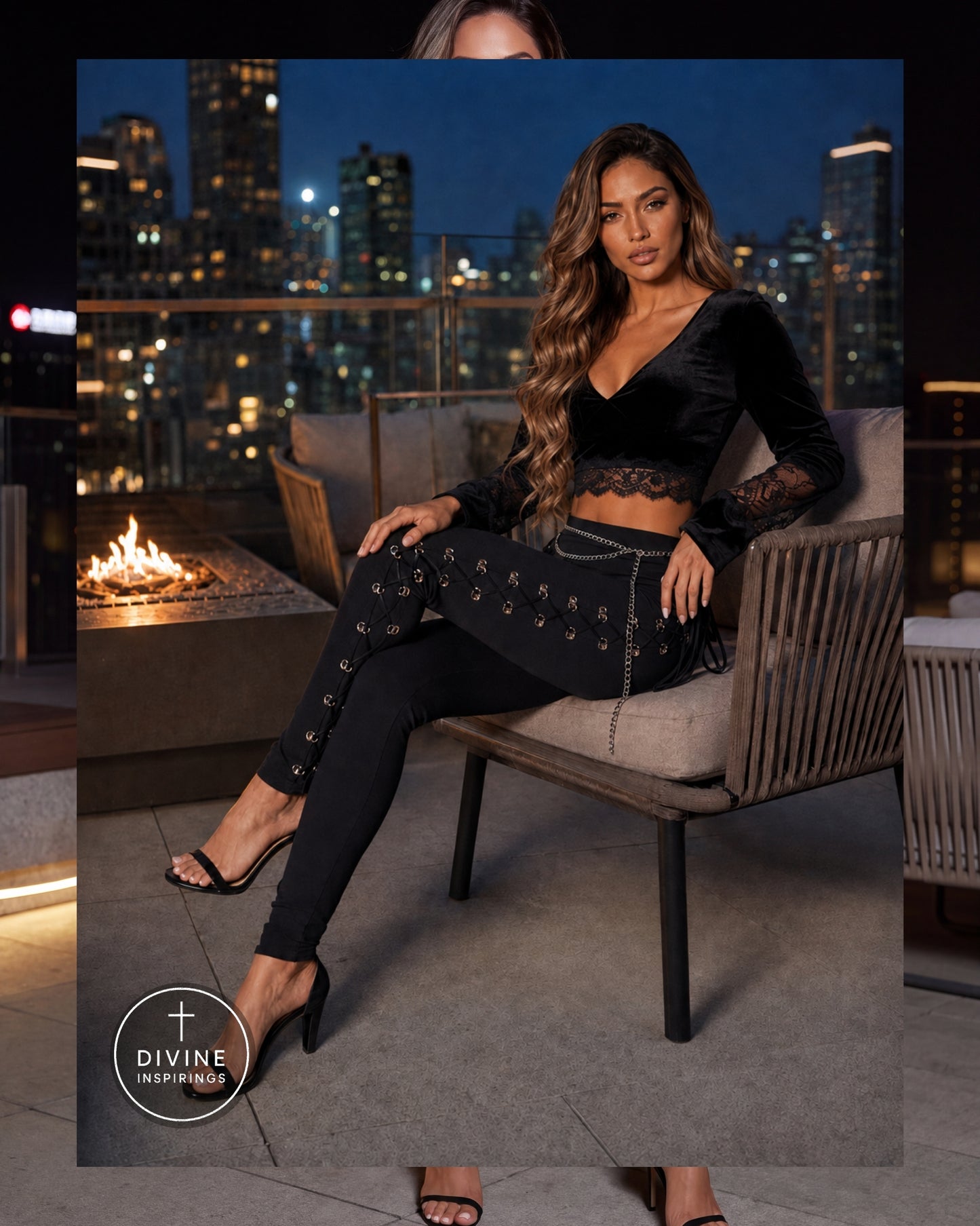 A woman models a black velvet long-sleeve crop top and matching embellished black leggings while sitting on a patio chair at night with a city skyline and fire pit in the background. A Divine Inspirings logo is visible in the lower left corner.