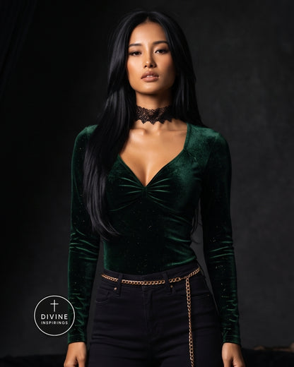 A woman modeling a dark green velvet long-sleeve top with a deep V-neck and gathered front, paired with black pants and a black lace choker, against a dark studio background. A Divine Inspirings logo is visible in the lower left corner.